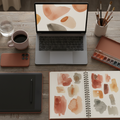 Laptop on a wooden desk with art supplies and a notebook displaying watercolor designs.